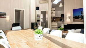 Ein moderner Essbereich mit Holztisch, weißen Stühlen und einer Zimmerpflanze. Im Hintergrund befindet sich eine Küche mit an der Wand montiertem Fernseher.