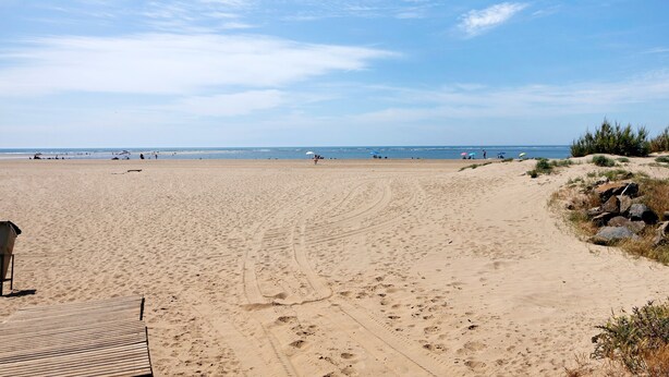 Una playa arenosa con huellas, una terraza de madera y vegetación escasa.