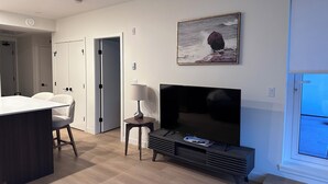 A modern living room with a flat-screen TV, a dining area with a table and chairs, and a painting of a rocky landscape. - Brand new condo in heart of Willoughby Town Centre Langley BC (Langley Township)