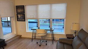 A living room with a sofa, a small table with a chair, a window with blinds, and a lamp. - Brand new condo in heart of Willoughby Town Centre Langley BC (Langley Township)