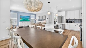 A modern kitchen with white cabinets, a dining area with a wooden table and white chairs, and a large chandelier. - Oceans East: Private heated pool, Free golf cart, 1 min beach, Game & Bunk rooms (Miramar Beach)