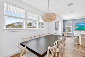A spacious dining area with a large table and chairs, large windows with blinds, a chandelier, and a television on the wall. - Oceans East: Private heated pool, Free golf cart, 1 min beach, Game & Bunk rooms (Miramar Beach)