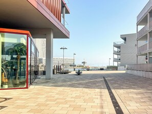 A modern building with a glass facade, a red-framed entrance, and a paved walkway leading to a waterfront. - Beach view vacation home 02 (Lübeck-Travemünde)
