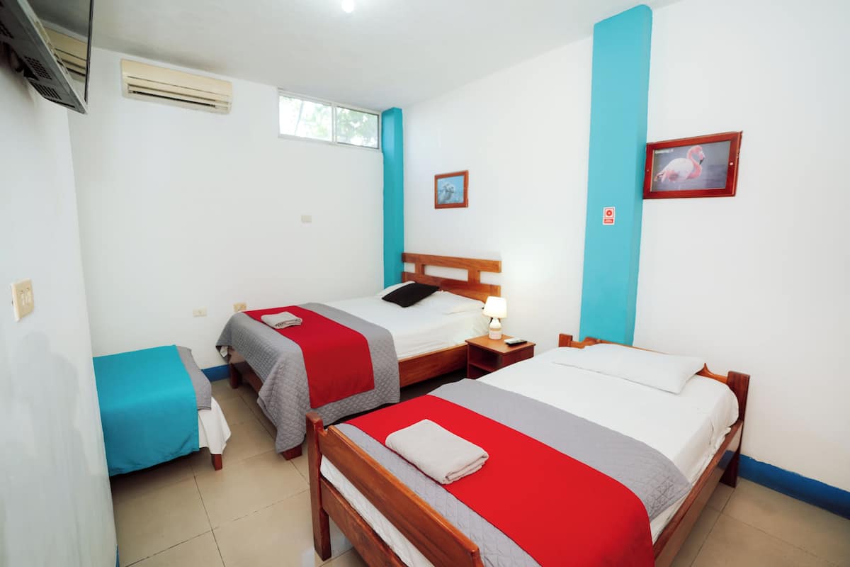 A hotel room with two single beds, a small table, a chair, an air conditioning unit, a window, and a wall-mounted picture.
