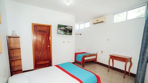 A room with two beds, a wooden desk, a wardrobe, a painting, and an air conditioning unit. - El Descanso del Petrel (Puerto Ayora)