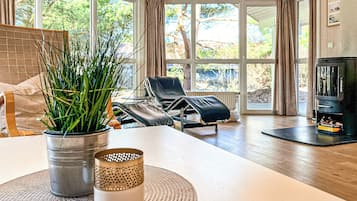 Ein modernes Wohnzimmer mit großem Fenster, Kamin, Lounge-Sessel und Esstisch mit Pflanze und Kerze.