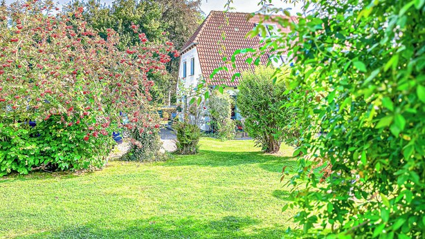 Et hus med rødt tegltag, omgivet af frodig vegetation og blomstrende buske.