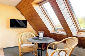 Une pièce chaleureuse avec un plafond en pente, un téléviseur fixé au mur, une table ronde avec des chaises et une fenêtre donnant sur un paysage verdoyant.