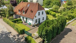 Et hvidt hus med rødt tegltag, omgivet af grøn vegetation og en belagt indkørsel.