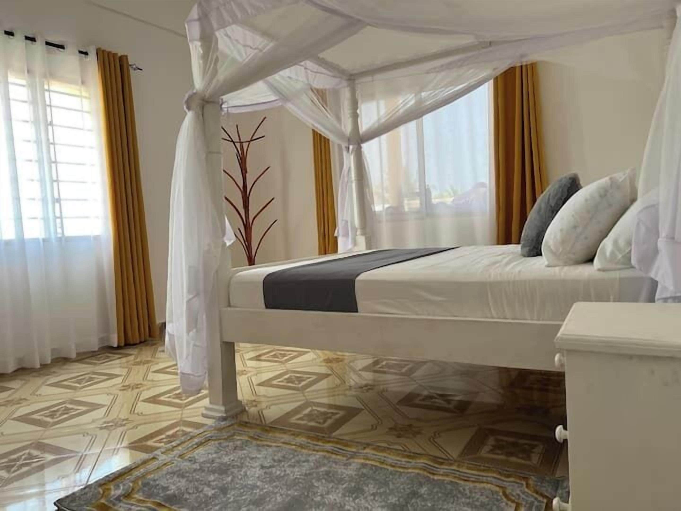 A bedroom with a canopy bed, a nightstand, a decorative plant, and patterned flooring.