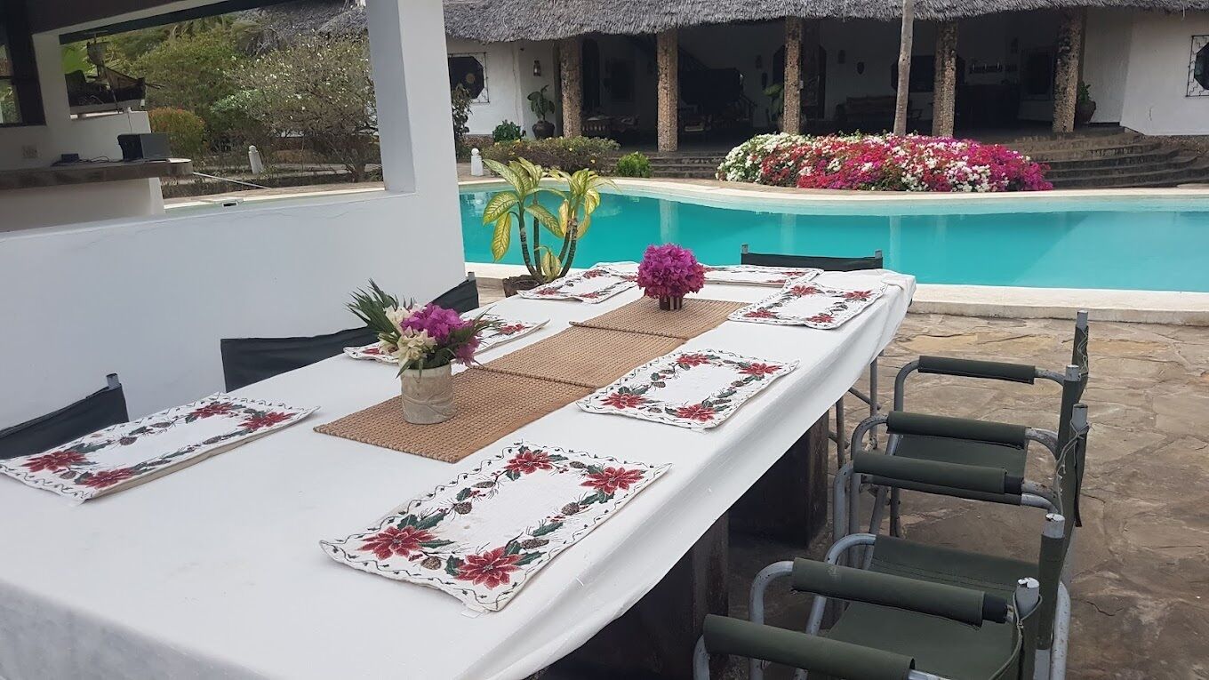 A dining table set with floral tablecloths and placemats, a vase with flowers, and a pool in the background.