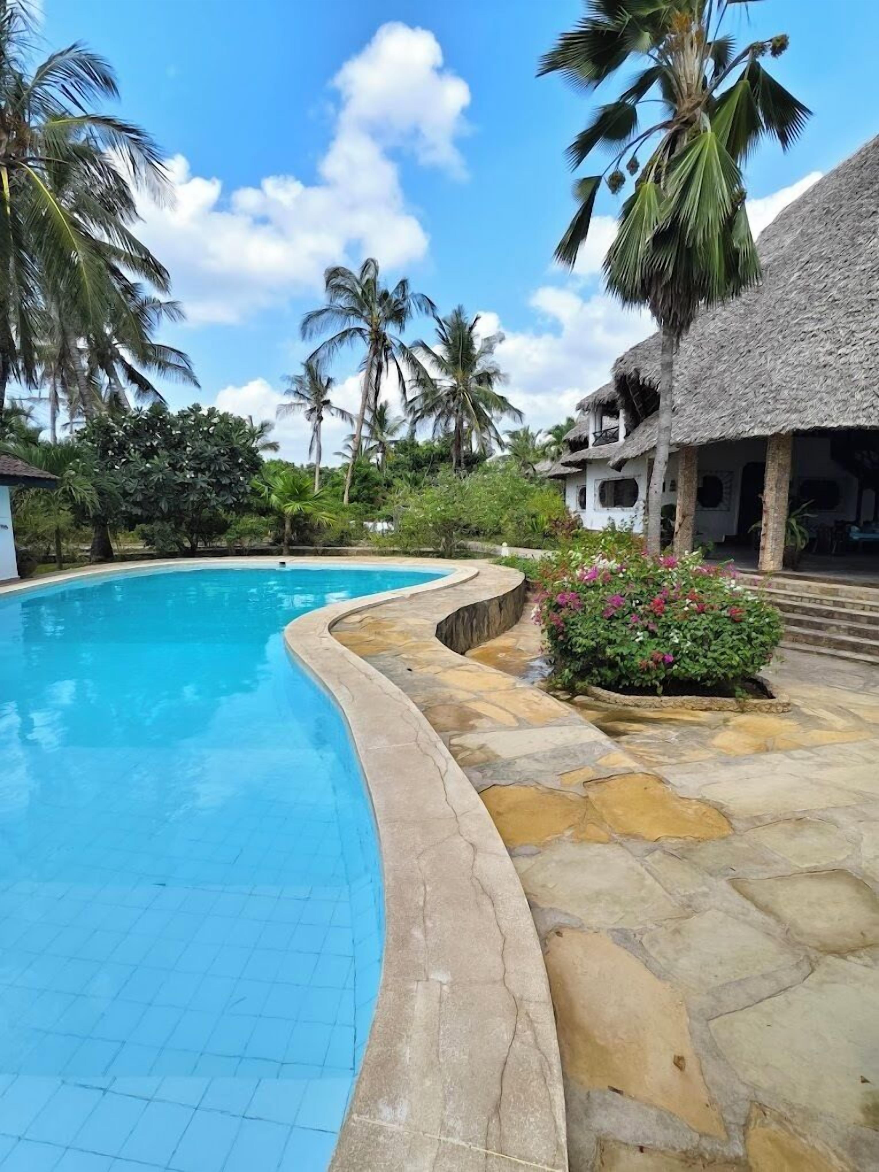 Una piscina con borde de piedra curvada, rodeada de vegetación tropical y un edificio con techo de paja.