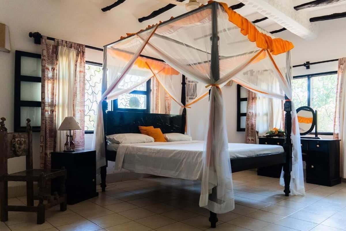 Un dormitorio con cama con dosel, mesita de noche, una silla y una ventana con cortinas.