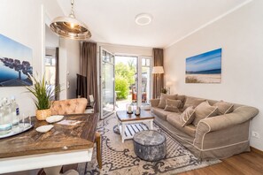 Een moderne woonkamer met een bank, een eettafel en een salontafel. Er is een groot raam met gordijnen, een schilderij aan de muur en een lamp.