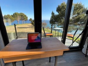 A laptop on a wooden table with a view of the sea and trees.