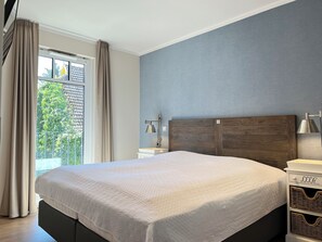 Ein modernes Schlafzimmer mit einem großen Bett, einem Nachttisch, einer Lampe und Blick ins Grüne.