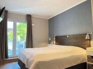 A hotel room with a large bed, a wooden headboard, a nightstand, a lamp, and a view of the outdoors. - Baltischer Hof 21 Apartment (Ostseebad Boltenhagen)