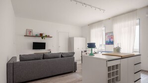 Ein modernes Wohnzimmer mit grauem Sofa, einem Flachbildfernseher, einem kleinen Couchtisch und einer Küchenzeile mit weißem Schrank und einem Fenster mit durchsichtigen Vorhängen.