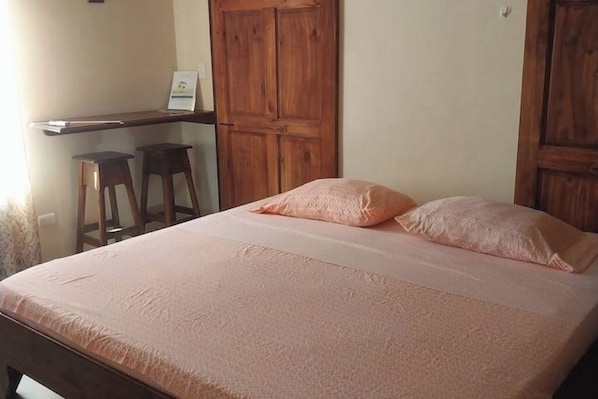 A bedroom with a bed, two wooden stools, a wooden door, and a small table with a sign.