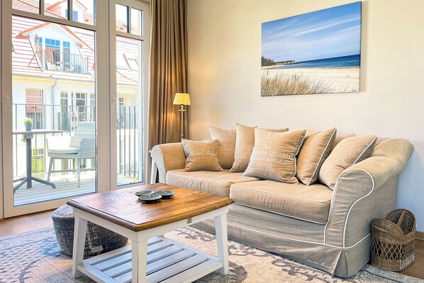 Ein gemütliches Wohnzimmer mit einem beigen Sofa, einem Holztisch und Blick auf ein Haus durch große Fenster.