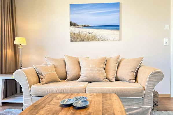 Un divano beige con cuscini coordinati, un tavolino in legno con piatti decorativi e una fotografia incorniciata di una spiaggia.