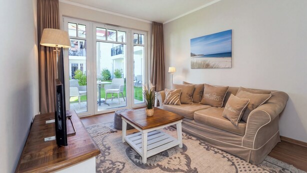 Ein Wohnzimmer mit einer Couch, einem Couchtisch, einem Fernseher auf einem Schrank und Blick auf einen Balkon mit Gartenmöbeln.