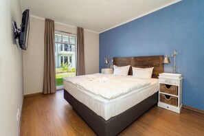 A hotel room with a large bed, a TV mounted on the wall, a nightstand with a lamp, and a view of the outdoors. - Baltischer Hof Apartment 09 (Ostseebad Boltenhagen)