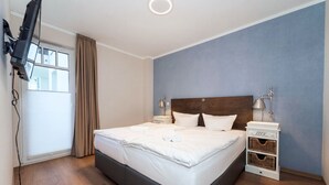 Ein Hotelzimmer mit einem Bett, einem an der Wand montierten Fernseher, einem Nachttisch mit Lampe und einem Fenster mit Vorhängen.