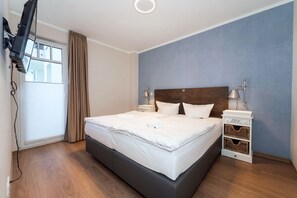 Ein Hotelzimmer mit einem Bett, einem an der Wand montierten Fernseher, einem Nachttisch mit Lampe und einem Fenster mit Vorhängen.