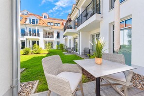 Une terrasse avec des meubles en osier et une table, une pelouse bien entretenue et, au fond, un bâtiment doté de plusieurs balcons.