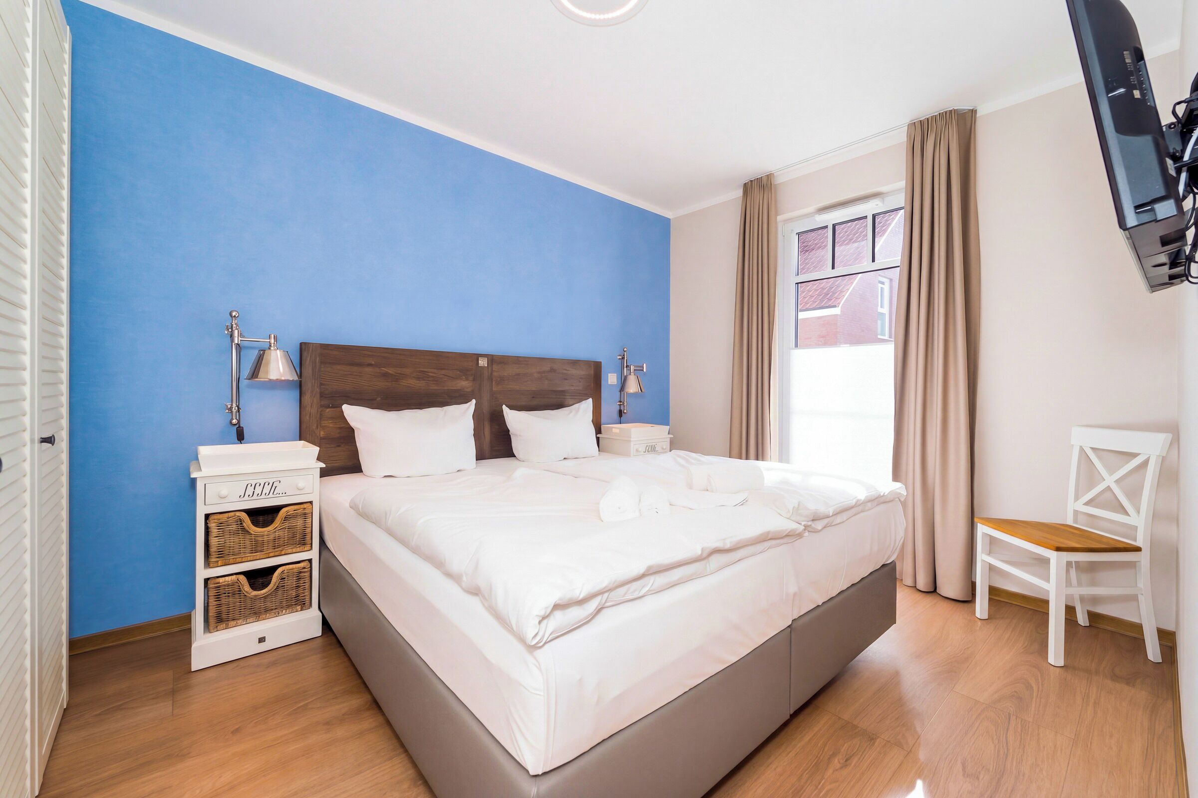 Un dormitorio con una cama grande, dos mesitas de noche, una silla, un televisor montado en la pared y una ventana con cortinas.