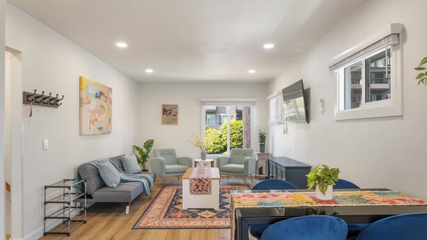 Uma sala de estar moderna com um sofá, cadeiras, uma mesa de centro e uma mesa de jantar com cadeiras. Há uma janela com persianas, uma TV fixada na parede e uma planta na mesa.