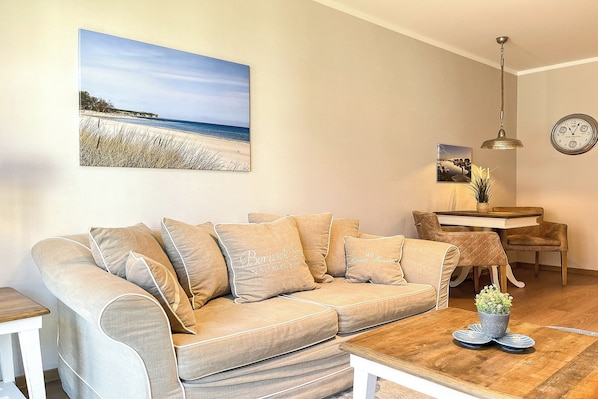Una sala de estar con un sofá color beige, una mesa de centro de madera con una planta en maceta, una mesa de comedor con sillas, un reloj de pared y una pintura de playa.