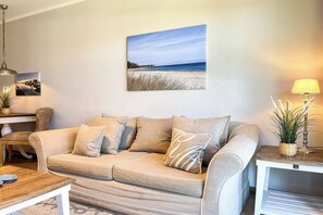 Ein Wohnzimmer mit einem beigen Sofa, einem Couchtisch, einer Lampe und einem Bild von einer Strandansicht.