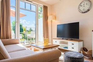 Ein Wohnzimmer mit einer Couch, einem Fernseher auf einem Holzständer, einer Lampe und Blick ins Freie durch große Fenster.