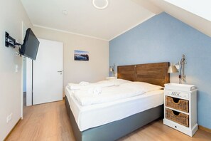 Ein Schlafzimmer mit einem Bett, einem an der Wand befestigten Fernseher, einem Nachttisch mit Lampe und einer kleinen Ablage mit Körben.