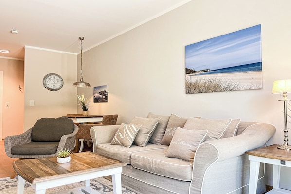 Ein Wohnzimmer mit einem beige-farbenen Sofa, einem Holztisch, einem Beistelltisch, einer Lampe, einer Uhr und einem Strandbild an der Wand.