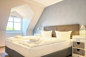 Ein ordentlich bezogenes Bett mit weißen Leinen und Handtüchern, ein hölzernes Kopfteil, ein Nachttisch mit Lampe und ein Fenster mit Tageslicht.
