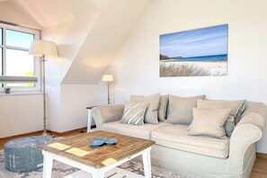 A cozy living room with a beige sofa, a wooden coffee table, and a painting of a beach scene. - Baltischer Hof 52 Apartment (Ostseebad Boltenhagen)
