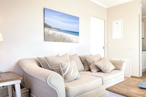 En beige sofa med flere puder, et træsidebord og et indrammet fotografi fra stranden på væggen.