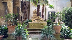 A courtyard with a statue of a reclining figure, surrounded by potted plants and steps leading up to a building with arched windows. - DUOMO, your private garden in TORRE: THE SFINGE OF THE SECRET GARDEN. (Firenze)