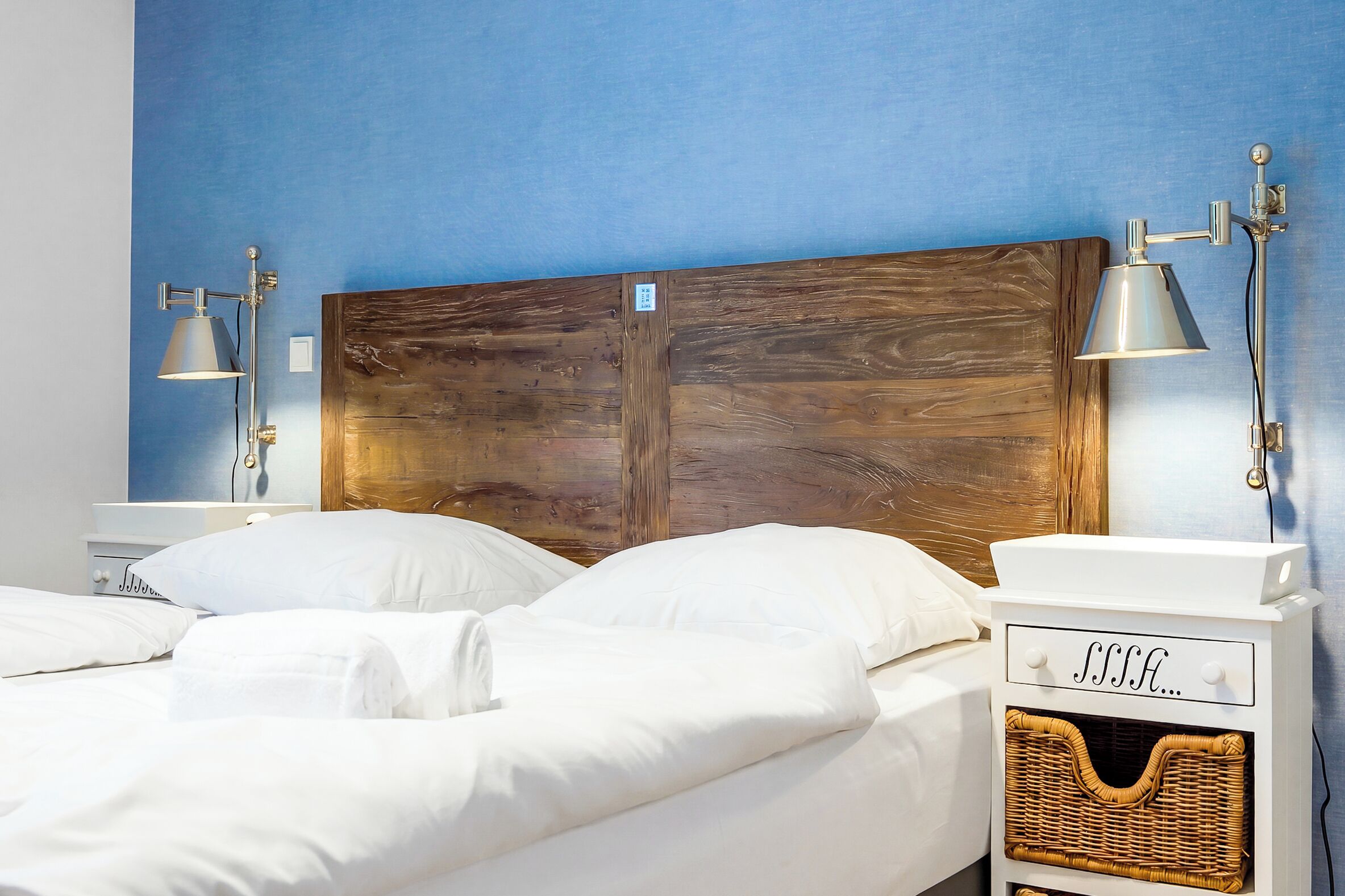 Een net opgemaakt bed met witte lakens, een houten hoofdeinde en twee bedlampjes.