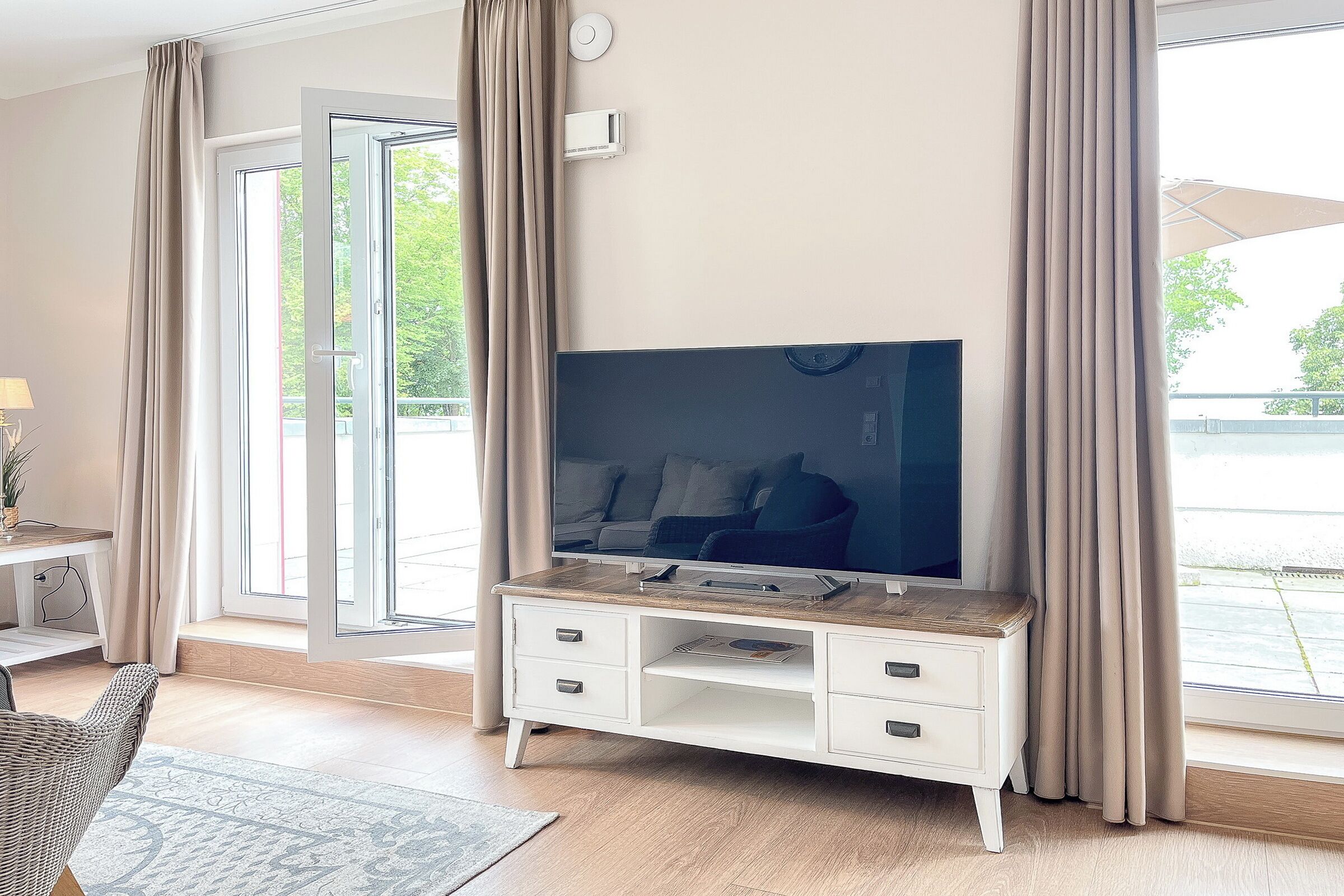 A modern living room with a flat-screen TV on a white console, large windows with curtains, a wicker chair, and a small table with a lamp.