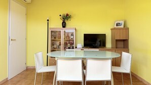 Un comedor con una mesa de vidrio y sillas blancas, un armario con puertas de cristal, un televisor y un mueble bajo con un cuadro encima.