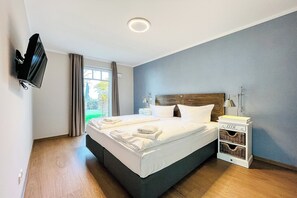 A modern bedroom with a large bed, a flat-screen TV mounted on the wall, a bedside table with a lamp, and a window with curtains. - Baltischer Hof apartment 18 (Ostseebad Boltenhagen)