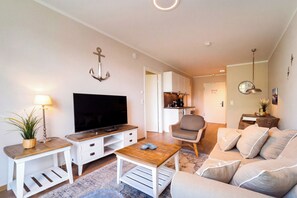 Ein Wohnzimmer mit Fernseher, Couchtisch, Sofa, Sessel und einer Küche im Hintergrund.