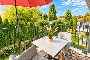 Ein Balkon mit Tisch und Stühlen, einem roten Sonnenschirm und Blick auf einen Garten mit verschiedenen Bäumen und einem Haus im Hintergrund.