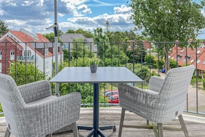 Ein Balkon mit Tisch und zwei Stühlen mit Blick auf eine Wohngegend, mit Häusern und einem roten Auto im Hintergrund.