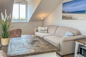 Ein Wohnzimmer mit einem beigefarbenen Sofa, einem Holztisch, einer Stehlampe und einem Gemälde mit einer Strandansicht.