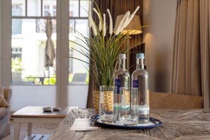 Ein Tisch mit Tablett, darauf drei Flaschen und zwei Gläser, eine Topfpflanze mit weißen Blüten und im Hintergrund ein Fenster mit Vorhängen.